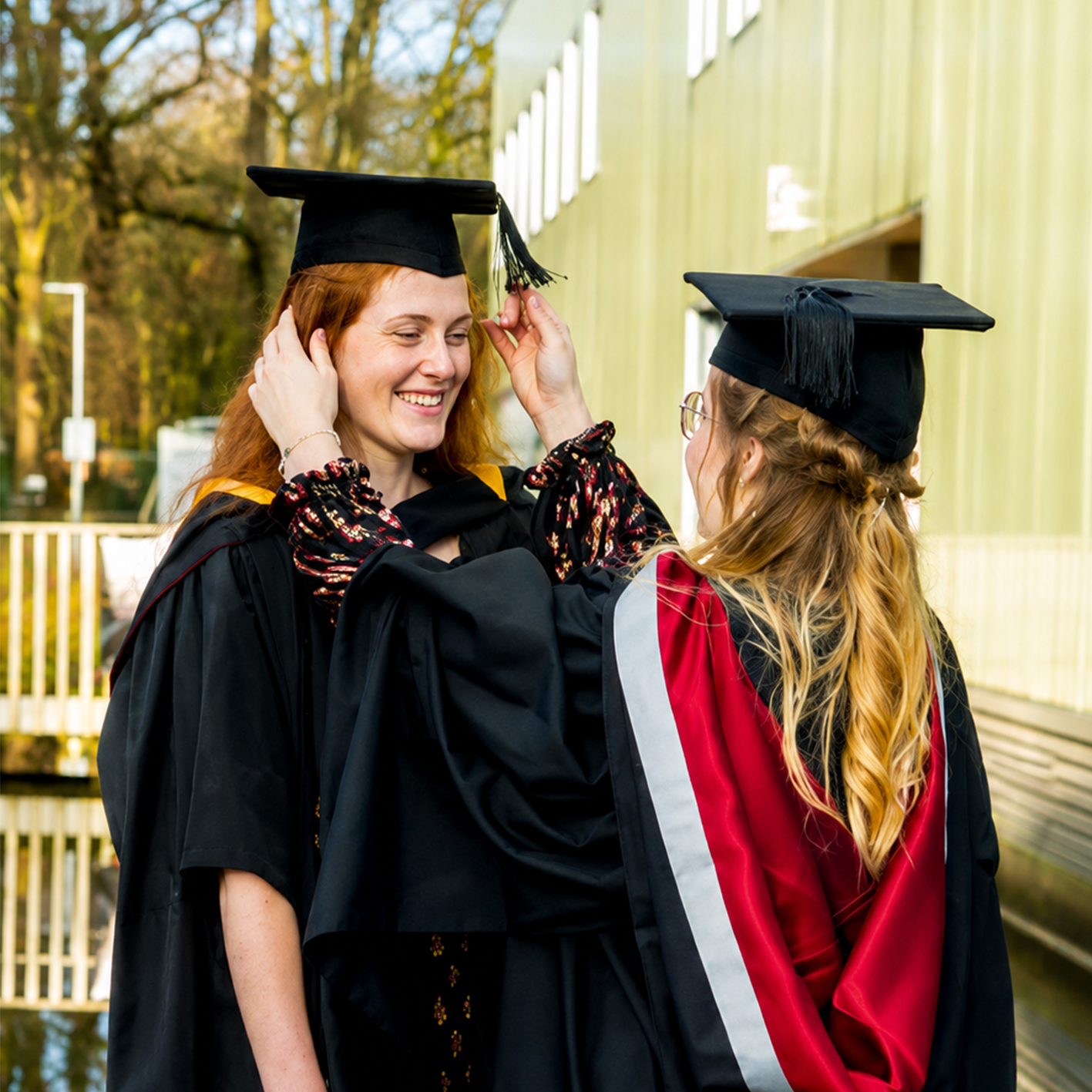 Two igirls in graduation gowns and caps outdoors, one adjusting the other's cap.